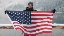Traveller Man Holding The Flag Of America Standing In The Snow-covered Mountains Neat Beautuful Lake. He Looking Into The Camera And Smiling. Hiker Traveller