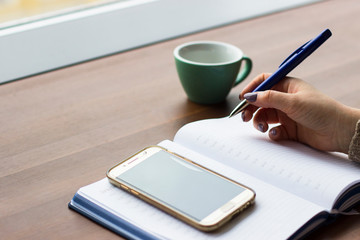 Close up of woman's hand writing something in a notebook on the background of a Cup, a phone and a wooden table. An unrecognized business woman is planning her day. Planning, work, business concept.