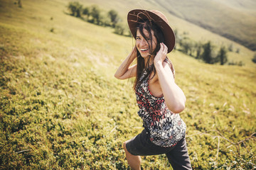 Stylish hipster girl in hat traveling on top of sunny mountains and smiling. Portrait of happy young woman with beautiful hair exploring mountains. Travel and wanderlust concept.