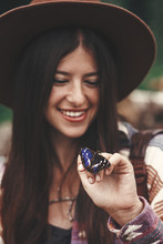 Stylish Hipster Girl In Hat Holding Purple Emperor Butterfly On Hand In Summer Mountains. Young Woman Traveler With Butterfly, Exploring Outdoors. Apatura Iris