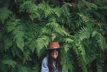 Stylish Hipster Girl In Hat Posing In Big Fern Leaves In Forest. Portrait Of Calm Young Woman In Green Leaves In Woods. Travel And Wanderlust Concept. Atmospheric Moment