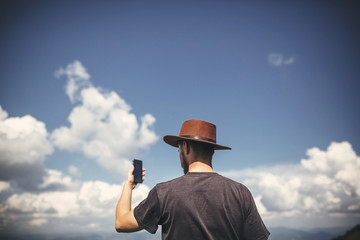 Wall Mural - Stylish hipster traveler  holding phone, standing on top of hills on  background of summer mountains. Copy space. Travel and wanderlust concept.