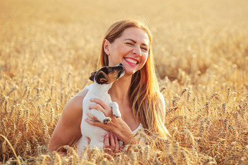 Young woman holds Jack Russell terrier puppy on her hands, laughing, dog is licking her cheeks and chin, sunset lit wheat field in background.