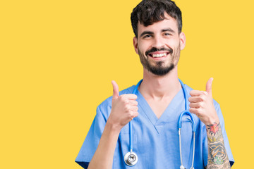 Sticker - Young handsome nurse man wearing surgeon uniform over isolated background success sign doing positive gesture with hand, thumbs up smiling and happy. Looking at the camera with cheerful expression