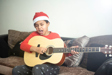 Cool mixed boy Matiss playing on guitar and singing a Christmas song. Boy in Santa Claus hat singing at his house on Happy New Year. Childrens hobby. Boy learning how to play guitar. 