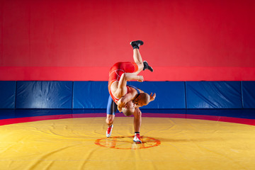 Two young men in blue and red wrestling tights are wrestlng and making a hip throw on a yellow wrestling carpet in the gym. The concept of fair wrestling