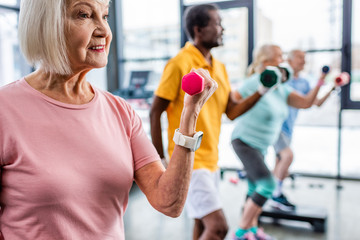 selective focus of senior sportswoman and her friends exercising with dumbbells at gym