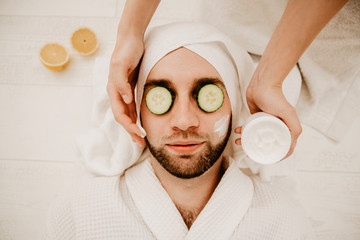 Relaxed bearded young man in a towel on his head being in spa salon with moisturizer cucumber and mask on the face