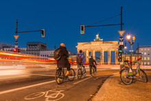 Backside Of The Brandenburg Gate At Night. Cyclists Are Waiting At The Traffic Light. Passing Cars In Long-term Exposure. An E-bike Is On The Roadside