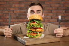 Young Hungry Man With Cutlery Eating Huge Burger At Table