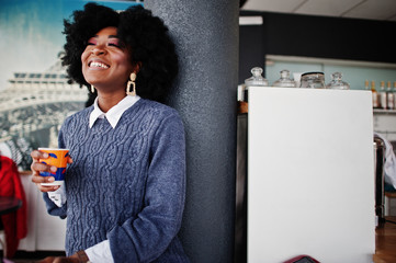 Curly hair african american woman wear on sweater posed at cafe indoor with cup of tea or coffee.