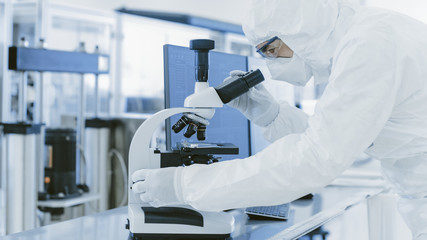 In Laboratory Scientist in Protective Clothes Doing Research Uses Microscope and Personal Computer. Modern Manufactory Producing Semiconductors and Pharmaceutical Items.