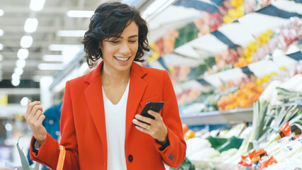 At the Supermarket: Portrait of a Beautiful Happy Young Woman Uses Smartphone While Standing at the Fresh Produce Section of the Store.