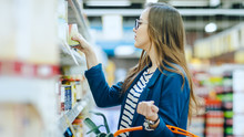 At The Supermarket: Beautiful Young Woman Browses Through The Canned Goods Section Of The Store. She Has Shopping Basket Full Of Healthy Food Items.