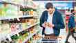 © Gorodenkoff - At the Supermarket: Handsome Man Uses Smartphone and Looks at Nutritional Value of the Canned Goods. He's Standing with Shopping Cart in Canned Goods Section.
