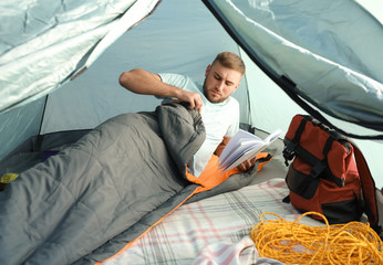 Wall Mural - Young man reading book in sleeping bag inside of camping tent