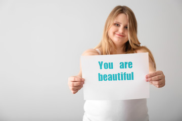 Wall Mural - Beautiful plus size girl holding paper sheet with text YOU ARE BEAUTIFUL on light background. Concept of body positivity