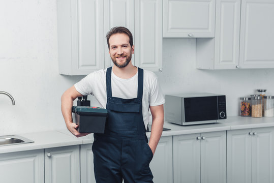 smiling bearded repairman in working overall holding toolbox in kitchen at home