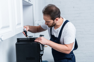 Wall Mural - bearded handyman in working overall repairing coffee machine by screwdriver in kitchen