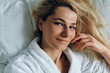 © iuricazac - Smiling blonde attractive young woman lying on the bed at home after bath in white bathrobe relaxing in the weekend. Beautiful female smiling and lookling to the camera on the bed in the morning.