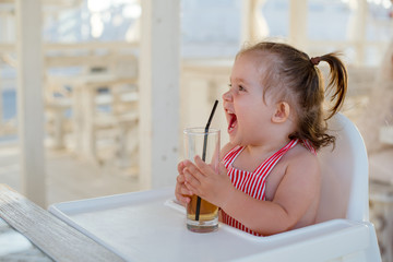 A little smiling toddler girl is sitting at a table in a summer cafe in the seaside resort.