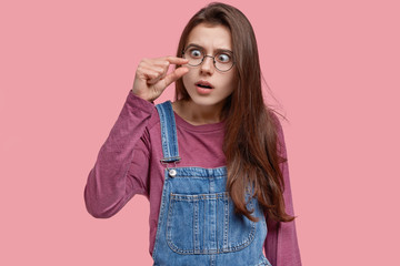 Wall Mural - Young woman shows small amount of something, demonstrates unimpressive size, shapes tiny thing, has scared expression, dressed in stylish clothes, models against pink studio wall. Little object