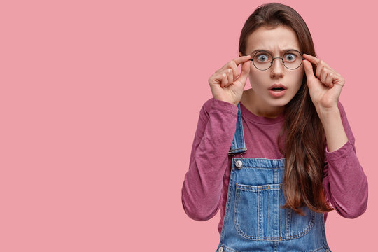 Photo of frightened young European woman keeps hands on frame of spectacles, has surprised expression, long hair, stares astonished, models over pink wall with copy space for your advertisement