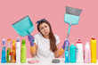 Dejected upset young housemaid frowns face in discontent, holds scoop and broom, does cleaning, wears headband and t shirt, isolated over pink background. Domestic routine and busyness concept