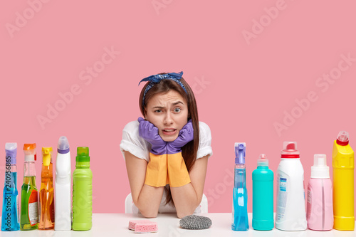 Fotografía Photo of attractive woman holds chin, frowns face, wears headband, protective rubber gloves, surrounded with cleaning accessories, poses at desk, feels tired of housework