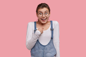 Wall Mural - Positive Caucasian woman points at herself, looks joyfully at camera, asks whether she is real candidate for winning contest, wears denim sarafan, round spectacles, models indoor, has toothy smile