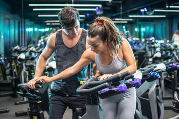 Wall Mural - Young attractive woman with handsome trainer setting up exercise bike before cycling workout in gym 