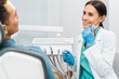 © LIGHTFIELD STUDIOS - cheerful female dentist holding mask and drill and smiling near patient