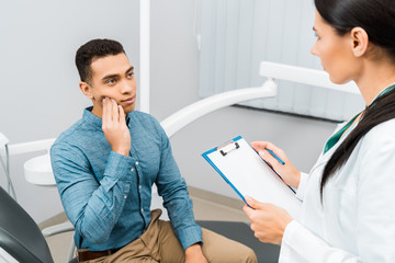 Wall Mural -  female dentist standing with clipboard and looking at handsome african american patient having toothache