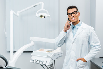 Wall Mural - smiling african american doctor in glasses and white coat talking on smartphone with hand in pocket