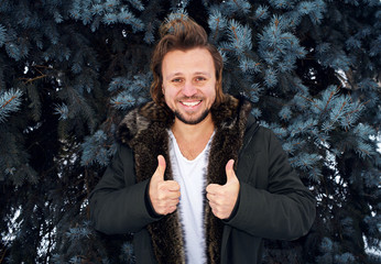 Young cheerful man showing thumbs up in front of the tree in winter.