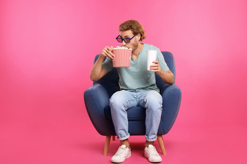 Man with 3D glasses, popcorn and beverage sitting in armchair during cinema show on color background