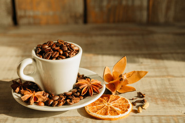 Cup with coffee beans and dried orange placed with star anise on wooden table lit by morning sunlight in cafe 