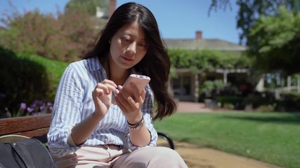 quick movement young confident businesswoman texting message on cellphone outdoors. asian lady sitting on bench in the green garden in summer using technology. house surrounding by trees and plants.