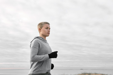 Picture of active healthy blonde short haired female athlete in gloves and hoodie enjoying morning cardio training, having thoughtful look. Pretty girl in sportswear running on cloudy summer day