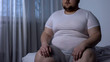 Fat man sitting on bed, weight gain due to sedentary lifestyle, hormonal disease