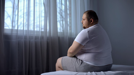 Depressed fat man sitting on bed at home, worried about overweight, insecurities