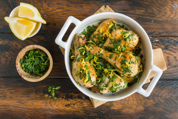 Baked chicken legs with fresh parsley and lemon juice and zest in white casserole on wooden rustic table, top view