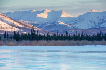 Snowy mountains in the rays of dawn. Winter landscape with the mountains and the frozen lake in Yakutia, Siberia at sunrise. Clear blue ice of the frozen lake at morning Frosty morning, day or evening