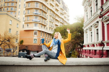 Stylish happy young blond woman in bright yellow coat holds coffee to go. Walking on the street. Portrait of a young cheerful woman at work break. Lifestyle concept 