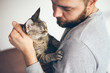 © veera - Close-up of beard man hugging cuddly cat, lifestyle portrait. Caring man is holding a Devon Rex -tabby kitty, both looking at each other with affection. Lifestyle, home interior photo
