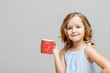© Olga - Portrait of a happy smiling little blonde girl on a gray background. The child holds a red mug