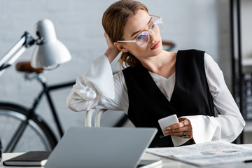 businesswoman in formal wear and glasses sitting at computer desk and using smartphone at workplace