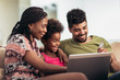 © Mediteraneo - Cute little Afro-American girl and her beautiful young parents using a laptop and doing shopping online while sitting on a sofa at home.