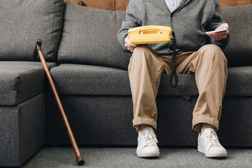 Wall Mural - cropped view of retired man holding smartphone and retro phone while sitting on sofa