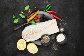 Cod fillets on a black ceramic plate for the preparation of a healthy dish with the addition of pink pepper, hot pepper, spinach, lemon, salt on a simple black background. Top view. Copy space